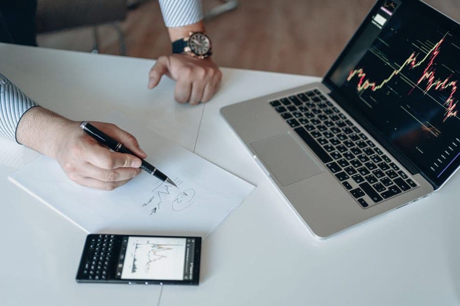Mão com caneta fazendo anotações e gráficos em papel, com notebook exibindo gráficos ao fundo