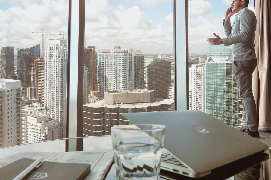 Executivo ao telefone diante de janela panorâmica com vista da cidade, mesa com notebook em primeiro plano